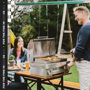 A man grilling outdoors while a woman sits at a table with drinks.